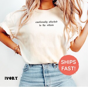 May include: Ivory-colored t-shirt with rolled sleeves and the phrase "emotionally attached to the villain" printed in black. The model is wearing blue jeans. The shirt is a casual, comfortable style.