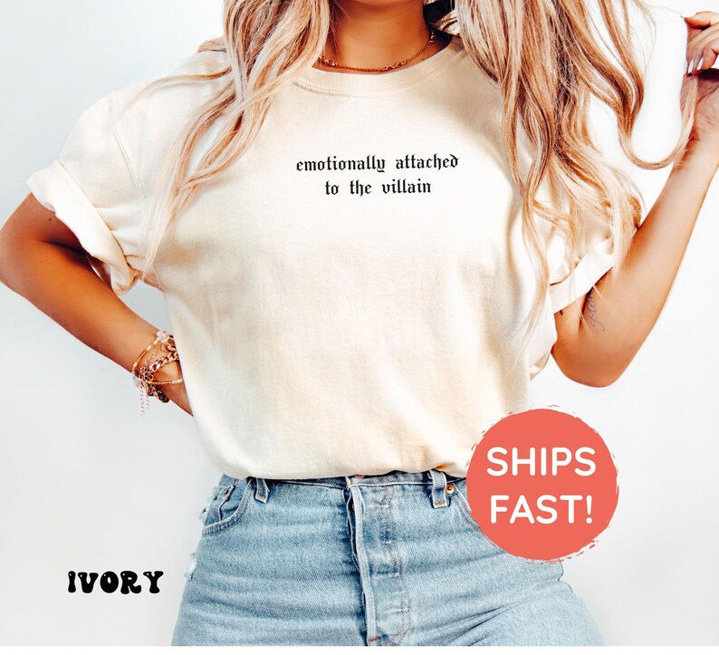 May include: Ivory-colored t-shirt with rolled sleeves and the phrase "emotionally attached to the villain" printed in black. The model is wearing blue jeans. The shirt is a casual, comfortable style.
