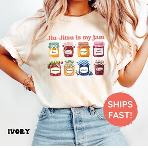 K&ouml;nnte beinhalten: Ein elfenbeinfarbenes T-Shirt mit dem Text "Jiu-Jitsu is my jam" &uuml;ber einer Grafik von acht bunten Marmeladengl&auml;sern. Jedes Glas ist mit einem anderen Jiu-Jitsu-Begriff beschriftet, darunter flow, base, triangle, half guard, armbar, mount, kimura und frame.