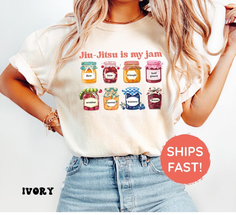 K&ouml;nnte beinhalten: Ein elfenbeinfarbenes T-Shirt mit dem Text "Jiu-Jitsu is my jam" &uuml;ber einer Grafik von acht bunten Marmeladengl&auml;sern. Jedes Glas ist mit einem anderen Jiu-Jitsu-Begriff beschriftet, darunter flow, base, triangle, half guard, armbar, mount, kimura und frame.