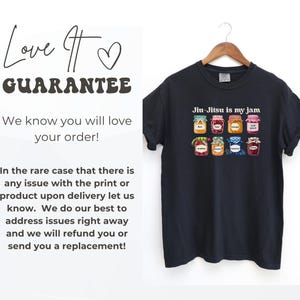 K&ouml;nnte beinhalten: Ein dunkelgraues Kurzarm-T-Shirt mit einem Grafikdruck von acht bunten Marmeladengl&auml;sern und dem Text "Jiu-Jitsu is my jam" dar&uuml;ber. Das T-Shirt h&auml;ngt auf einem Holzkleiderb&uuml;gel vor einem schlichten wei&szlig;en Hintergrund.