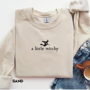 May include: A sand-colored crewneck sweatshirt featuring black embroidery of a witch flying on a broomstick above the text "a little witchy" centered on the chest. The garment has long sleeves and a ribbed collar, with the word "SAND" printed below.