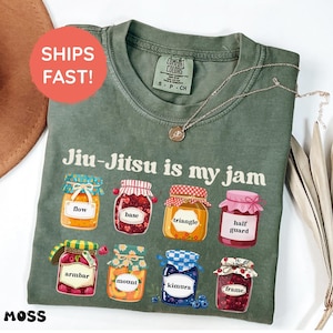 K&ouml;nnte beinhalten: Ein moosgr&uuml;nes Kurzarm-T-Shirt mit dem Text Jiu-Jitsu is my jam &uuml;ber acht illustrierten Marmeladengl&auml;sern. Jedes Glas ist mit einem anderen Begriff beschriftet: flow, base, triangle, half guard, armbar, mount, kimura und frame.