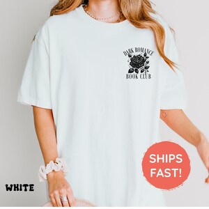 May include: A white short-sleeved t-shirt featuring a black graphic on the upper left chest. The graphic depicts a rose with leaves and the text "DARK ROMANCE BOOK CLUB" arched above and below the flower in a serif font style.
