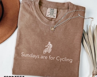May include: An espresso-colored crew neck t-shirt featuring a white graphic of a person riding a bicycle above the text "Sundays are for Cycling". The shirt has a Comfort Colors brand label on the inside collar and is made of 100% cotton fabric.