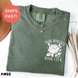 May include: A moss green short-sleeve t-shirt featuring a white graphic on the chest. The graphic depicts a rose surrounded by small stars and the text "DARK ROMANCE BOOK CLUB" in a serif font. The shirt has a crew neckline and a visible brand label.