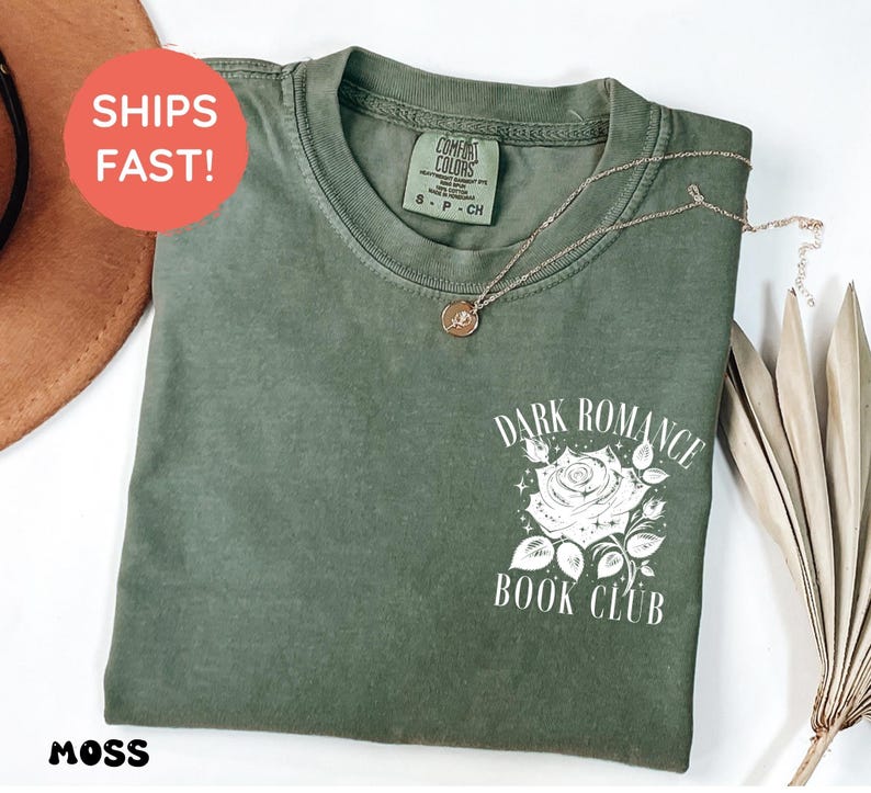 May include: A moss green short-sleeve t-shirt featuring a white graphic on the chest. The graphic depicts a rose surrounded by small stars and the text "DARK ROMANCE BOOK CLUB" in a serif font. The shirt has a crew neckline and a visible brand label.