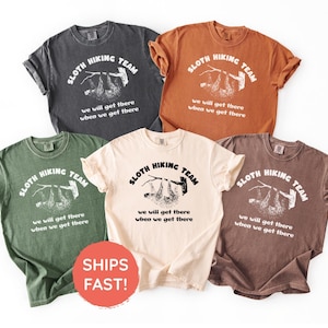 Könnte beinhalten: Fünf T-Shirts in verschiedenen Farben: Anthrazit, Rost, Olivgrün, Creme und Braun. Jedes Shirt zeigt eine Grafik eines Faultiers, das an einem Ast hängt, mit dem Text "SLOTH HIKING TEAM" und "we will get there when we get there."