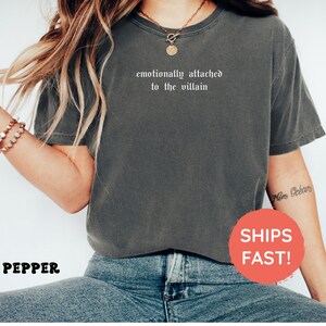 May include: A gray t-shirt with the text "emotionally attached to the villain" in white. The shirt is worn with blue jeans. A red circle with the words "SHIPS FAST!" is visible.