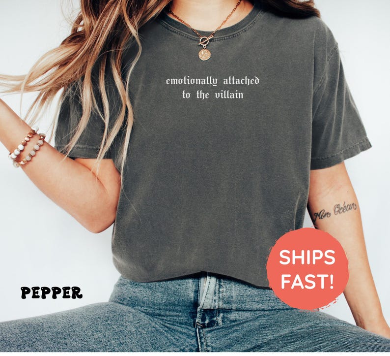 May include: A gray t-shirt with the text "emotionally attached to the villain" in white. The shirt is worn with blue jeans. A red circle with the words "SHIPS FAST!" is visible.