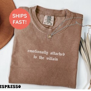 May include: Espresso-colored t-shirt with the phrase "emotionally attached to the villain" in white gothic font. A silver necklace with a pendant rests on the shirt. The shirt is folded and displayed on a white surface.