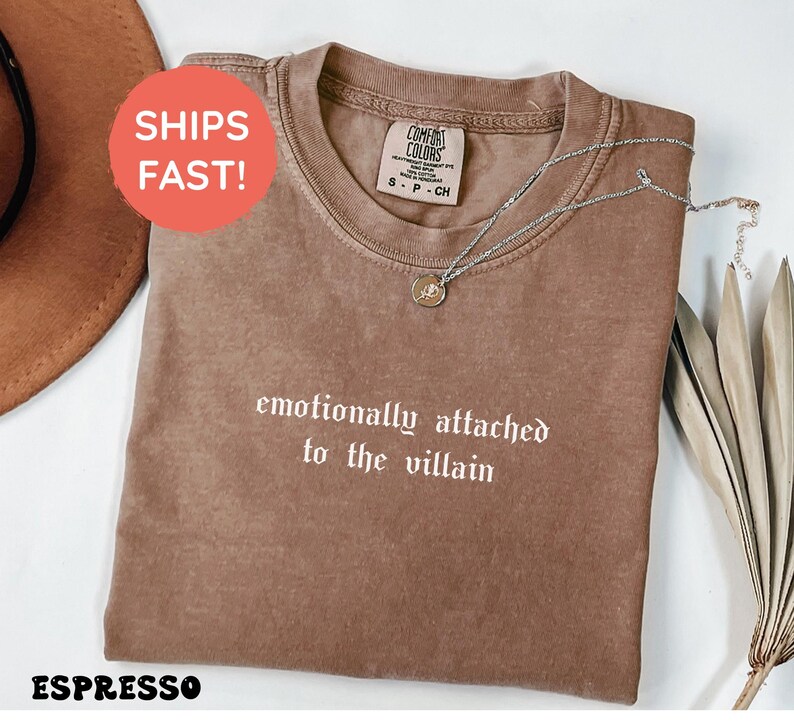 May include: Espresso-colored t-shirt with the phrase "emotionally attached to the villain" in white gothic font. A silver necklace with a pendant rests on the shirt. The shirt is folded and displayed on a white surface.