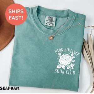 May include: A seafoam green short-sleeve t-shirt featuring a white graphic on the chest. The graphic depicts a rose surrounded by small stars and the text "DARK ROMANCE BOOK CLUB" in a serif font. The shirt has a crew neckline and a tag.