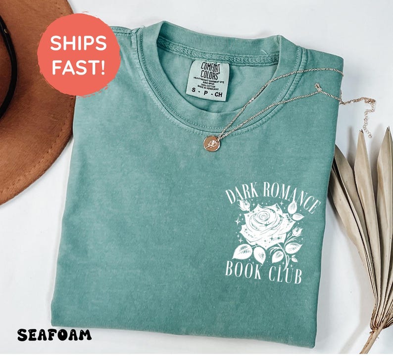 May include: A seafoam green short-sleeve t-shirt featuring a white graphic on the chest. The graphic depicts a rose surrounded by small stars and the text "DARK ROMANCE BOOK CLUB" in a serif font. The shirt has a crew neckline and a tag.