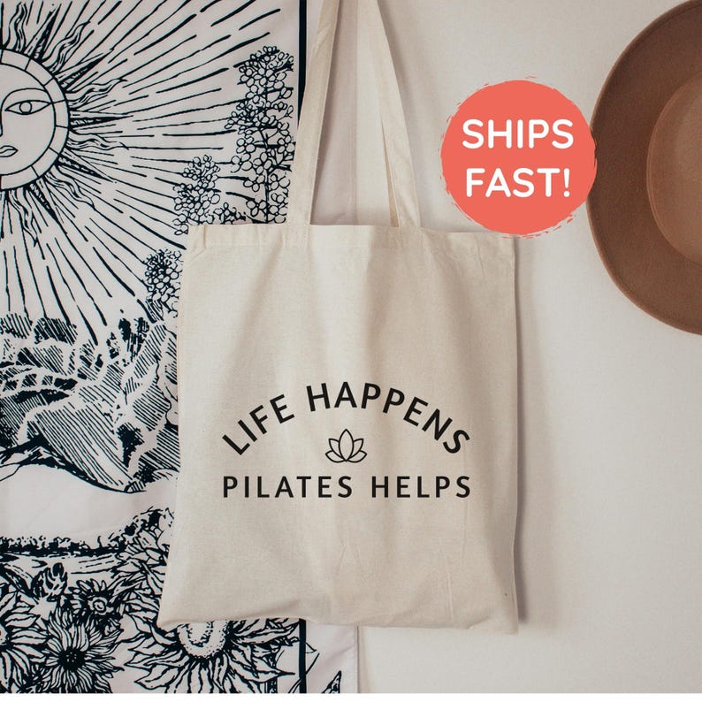 May include: A cream-colored canvas tote bag with two shoulder straps. The front of the bag features black text that reads "LIFE HAPPENS" in an arc, a small lotus flower icon in the center, and the text "PILATES HELPS" underneath.