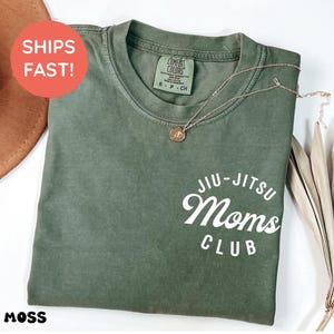Może przedstawiać: Koszulka z okrągłym dekoltem w kolorze mchu z białym napisem Jiu-Jitsu Moms Club nadrukowanym w prawym dolnym rogu. Koszulka posiada metkę marki Comfort Colors wewnątrz kołnierza i jest prezentowana ze złotym naszyjnikiem leżącym na tkaninie.