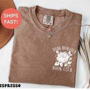 May include: A short-sleeve crewneck t-shirt in a solid espresso brown color. The front features a white graphic print of a rose with leaves and stars, accompanied by the text "DARK ROMANCE BOOK CLUB" in a clean, white serif font.