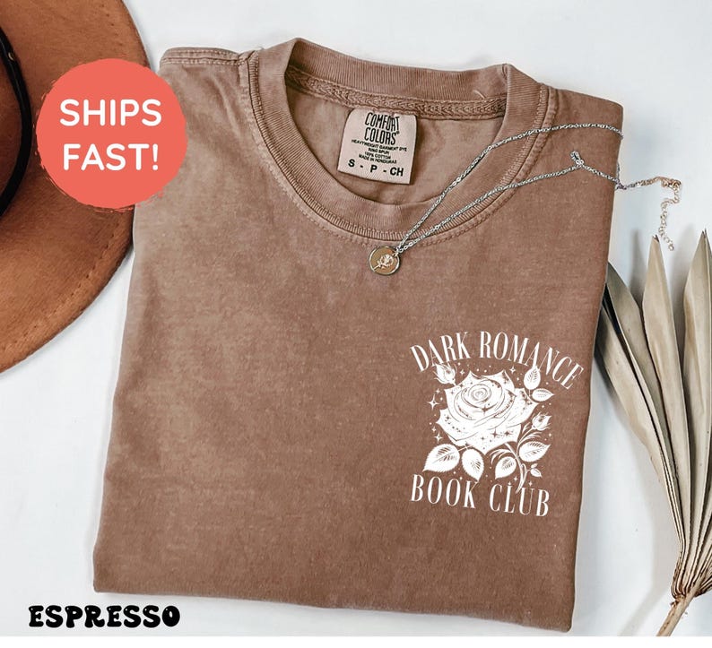 May include: A short-sleeve crewneck t-shirt in a solid espresso brown color. The front features a white graphic print of a rose with leaves and stars, accompanied by the text "DARK ROMANCE BOOK CLUB" in a clean, white serif font.