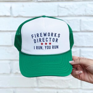 May include: Green and white trucker hat with the text "FIREWORKS DIRECTOR I RUN, YOU RUN" in navy blue. The hat has a green brim and mesh back, with three red stars below the word "DIRECTOR".