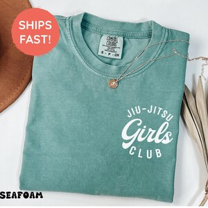Może przedstawiać: Zielona koszulka z napisem "Jiu-Jitsu Girls Club" w kolorze białym. Złoty naszyjnik z wisiorkiem jest przerzucony przez koszulkę. Koszulka ma okrągły dekolt i małą metkę. Czerwone koło z napisem "Ships Fast!" znajduje się po lewej stronie u g&oacute;ry.