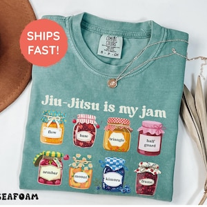 K&ouml;nnte beinhalten: Ein kurz&auml;rmeliges T-Shirt in Salbeigr&uuml;n mit dem Text Jiu-Jitsu is my jam und acht Abbildungen von Marmeladengl&auml;sern mit den Aufschriften flow, base, triangle, half guard, armbar, mount, kimura und frame. Das T-Shirt hat ein Comfort Colors Etikett.