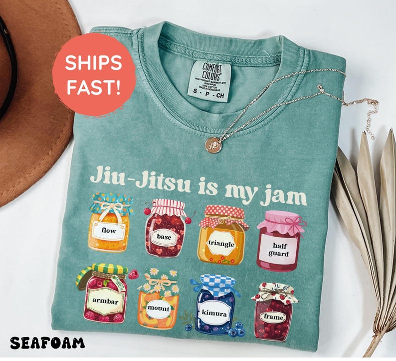 K&ouml;nnte beinhalten: Ein kurz&auml;rmeliges T-Shirt in Salbeigr&uuml;n mit dem Text Jiu-Jitsu is my jam und acht Abbildungen von Marmeladengl&auml;sern mit den Aufschriften flow, base, triangle, half guard, armbar, mount, kimura und frame. Das T-Shirt hat ein Comfort Colors Etikett.