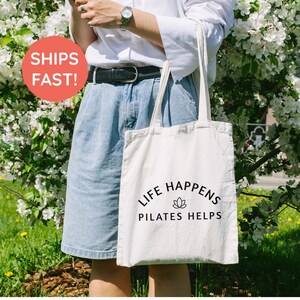 May include: A cream-colored canvas tote bag featuring the black printed text 'LIFE HAPPENS PILATES HELPS' with a small lotus flower icon centered between the two lines of text. The bag has two long shoulder straps and a rectangular shape.