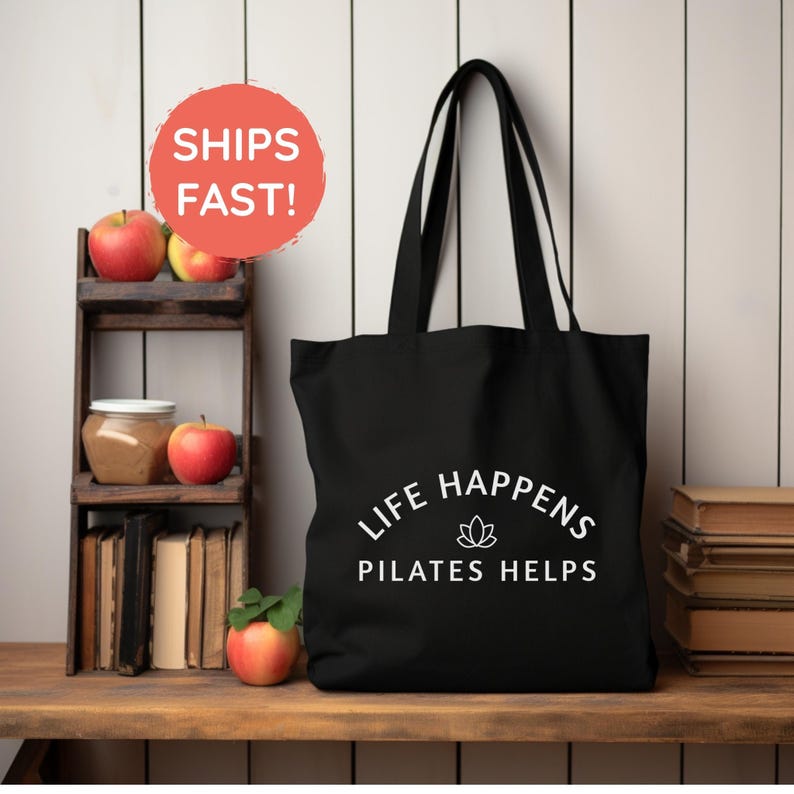May include: A black tote bag featuring the white text "LIFE HAPPENS PILATES HELPS" centered on the front, accompanied by a small white lotus flower icon positioned between the two lines of text.
