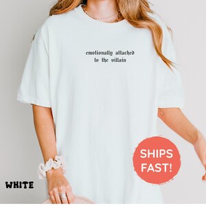 May include: White t-shirt with the text "emotionally attached to the villain" in black gothic font. The shirt is a relaxed fit and has short sleeves. The word "WHITE" is printed in a black font at the bottom left.