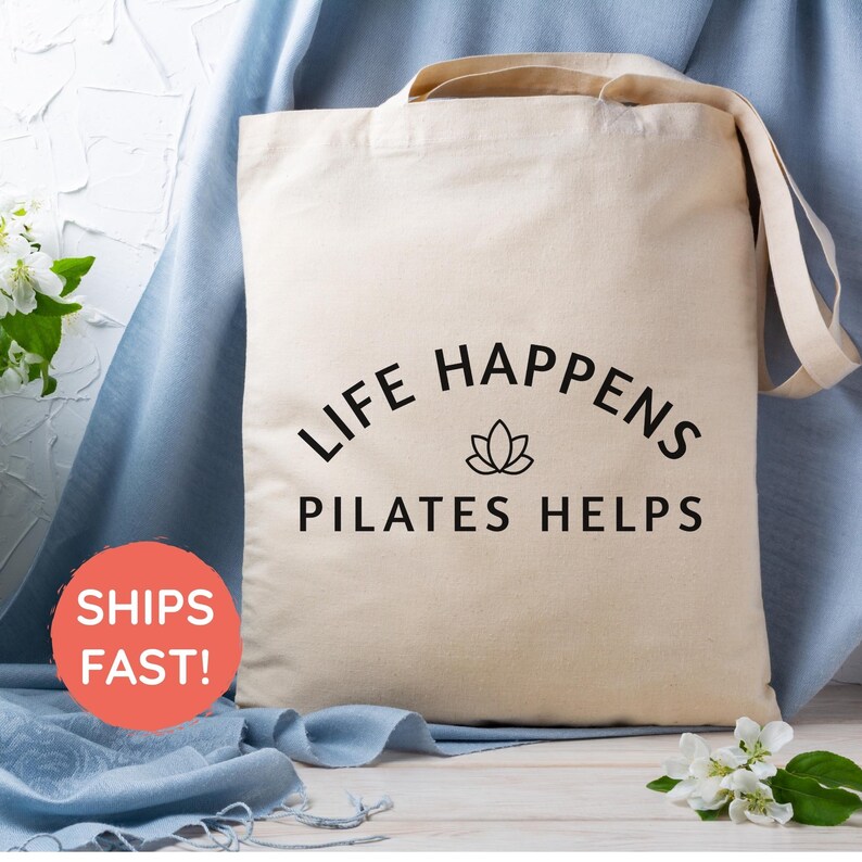 May include: A beige canvas tote bag featuring the black printed text LIFE HAPPENS PILATES HELPS with a small lotus flower icon centered between the two lines of text. The bag has two shoulder straps and a rectangular shape.