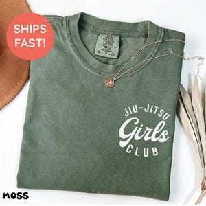 Może przedstawiać: Koszulka w kolorze mchu z białym napisem "Jiu-Jitsu Girls Club". Srebrny naszyjnik z wisiorkiem spoczywa na koszulce. Koszulka jest złożona i ma metkę Comfort Colors.