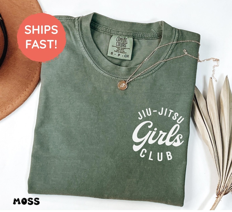 Może przedstawiać: Koszulka w kolorze mchu z białym napisem "Jiu-Jitsu Girls Club". Srebrny naszyjnik z wisiorkiem spoczywa na koszulce. Koszulka jest złożona i ma metkę Comfort Colors.