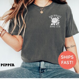 May include: A charcoal gray short-sleeve t-shirt featuring a white graphic on the left chest. The graphic depicts a rose illustration surrounded by the text "DARK ROMANCE BOOK CLUB". The shirt is paired with blue denim jeans.