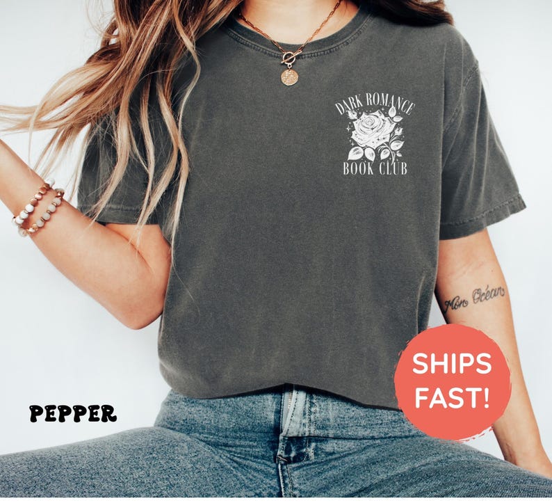May include: A charcoal gray short-sleeve t-shirt featuring a white graphic on the left chest. The graphic depicts a rose illustration surrounded by the text "DARK ROMANCE BOOK CLUB". The shirt is paired with blue denim jeans.
