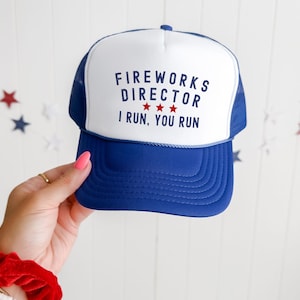 May include: A blue and white trucker hat with the text "FIREWORKS DIRECTOR" and "I RUN, YOU RUN" in navy blue. The hat has a blue brim and mesh backing. Three red stars are between the words "DIRECTOR" and "I".