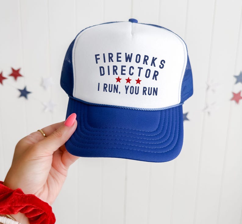 May include: A blue and white trucker hat with the text "FIREWORKS DIRECTOR" and "I RUN, YOU RUN" in navy blue. The hat has a blue brim and mesh backing. Three red stars are between the words "DIRECTOR" and "I".