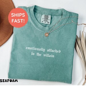 May include: Seafoam green t-shirt with the phrase "emotionally attached to the villain" in white script. A gold-colored necklace with a pendant rests on the shirt. The shirt is a Comfort Colors brand.