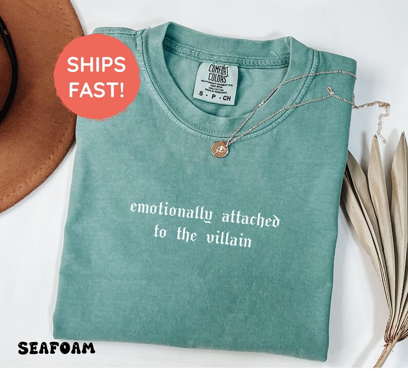 May include: Seafoam green t-shirt with the phrase "emotionally attached to the villain" in white script. A gold-colored necklace with a pendant rests on the shirt. The shirt is a Comfort Colors brand.
