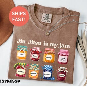 K&ouml;nnte beinhalten: Ein braunes kurz&auml;rmeliges T-Shirt mit dem Text Jiu-Jitsu is my jam und acht Illustrationen von Marmeladengl&auml;sern, beschriftet mit flow, base, triangle, half guard, armbar, mount, kimura und frame. Das T-Shirt hat ein Comfort Colors Etikett.