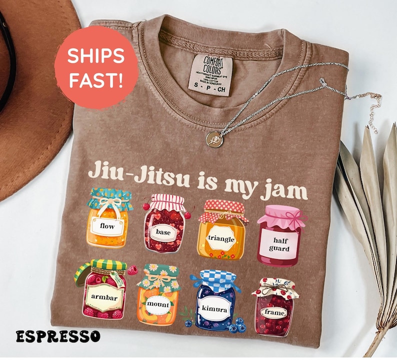 K&ouml;nnte beinhalten: Ein braunes kurz&auml;rmeliges T-Shirt mit dem Text Jiu-Jitsu is my jam und acht Illustrationen von Marmeladengl&auml;sern, beschriftet mit flow, base, triangle, half guard, armbar, mount, kimura und frame. Das T-Shirt hat ein Comfort Colors Etikett.
