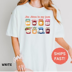 K&ouml;nnte beinhalten: Ein wei&szlig;es T-Shirt mit einer Grafik von acht bunten Gl&auml;sern, die mit verschiedenen Fr&uuml;chten und Beeren gef&uuml;llt sind. Jedes Glas ist mit Text beschriftet, darunter flow, base, triangle, half guard, armbar, mount, kimura und frame. Der Text Jiu-Jitsu is my jam ist in Rot &uuml;ber den Gl&auml;sern gedruckt.