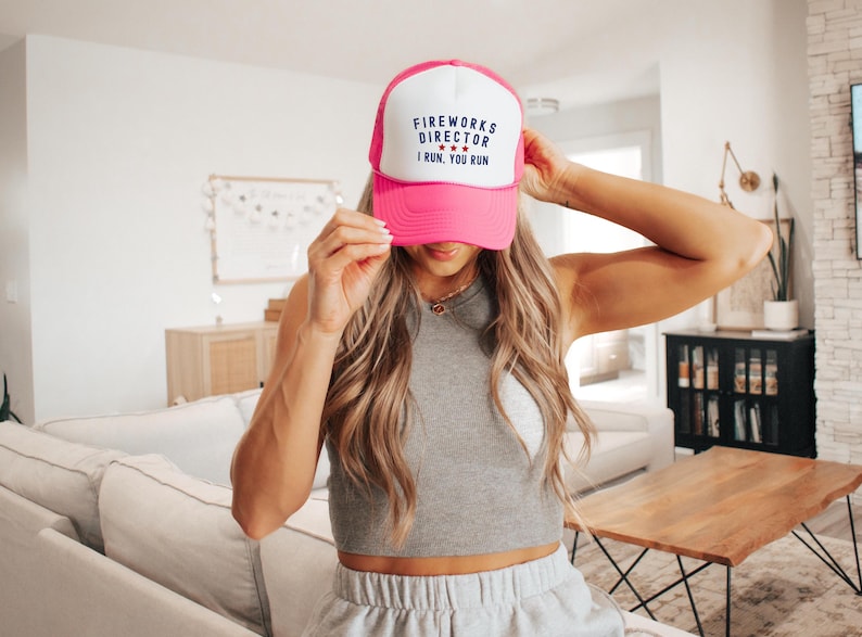 May include: A pink and white trucker hat with the text "FIREWORKS DIRECTOR I RUN, YOU RUN" in black and blue. The hat has a curved brim and a mesh back. The hat is a fun accessory for Independence Day celebrations.