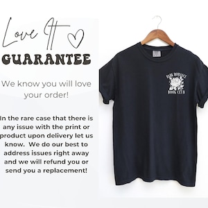 May include: A black short-sleeve t-shirt hanging on a wooden hanger. The shirt features a white graphic on the left chest area depicting a rose with the text "DARK ROMANCE BOOK CLUB" arched above and below the flower in a serif font.