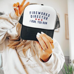 May include: A black and white trucker hat with the text "FIREWORKS DIRECTOR" in blue, three red stars, and the text "I RUN, YOU RUN". The hat has a white front panel and a black mesh back.