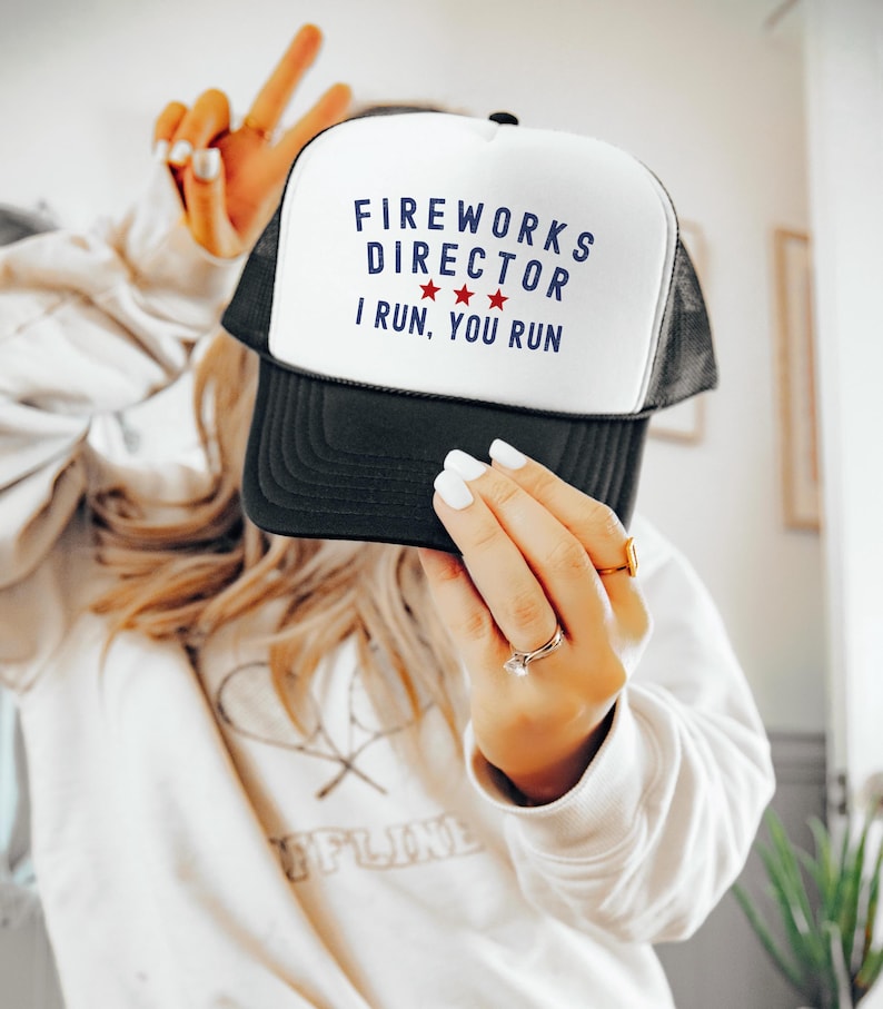 May include: A black and white trucker hat with the text "FIREWORKS DIRECTOR" in blue, three red stars, and the text "I RUN, YOU RUN". The hat has a white front panel and a black mesh back.
