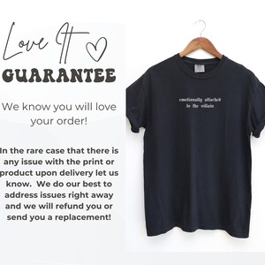 May include: Black t-shirt with the phrase "emotionally attached to the villain" in white lettering. The shirt is hanging on a wooden hanger. Text on the left side reads "Love It Guarantee" and a customer satisfaction statement.