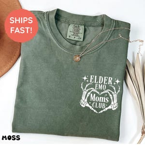 May include: A moss green t-shirt with a crew neck and short sleeves. The shirt has a white graphic of skeleton hands forming a heart with the text "ELDER EMO MOMS CLUB". A gold necklace with a pendant is on the shirt.