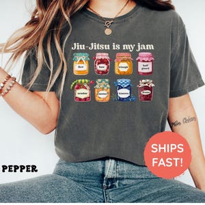K&ouml;nnte beinhalten: Ein anthrazitfarbenes T-Shirt mit dem Text Jiu-Jitsu is my jam &uuml;ber einer Grafik von acht bunten Gl&auml;sern. Jedes Glas ist mit einem bestimmten Begriff beschriftet: flow, base, triangle, half guard, armbar, mount, kimora und frame.