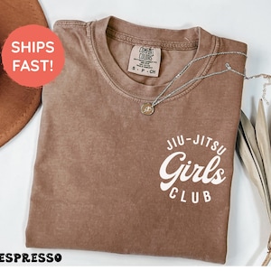 Może przedstawiać: Koszulka w kolorze espresso z białym napisem "Jiu-Jitsu Girls Club". Koszulka ma okrągły dekolt i małą metkę na kołnierzu. Srebrny naszyjnik z wisiorkiem jest zarzucony na koszulkę.