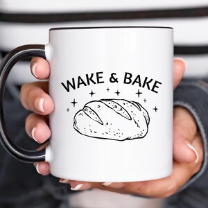 Peut inclure: Mug en céramique blanche avec une anse et un bord noirs, avec le texte "WAKE & BAKE" au-dessus d'une illustration de pain. Le design est en noir et blanc, avec des étoiles. Le mug est tenu par une personne.